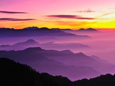 the sun is setting over some mountains in the foggy mist filled sky, with purple and yellow hues