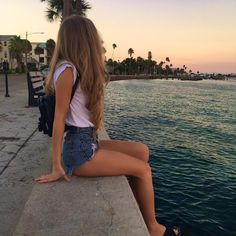 a woman sitting on the edge of a pier looking at the water and palm trees