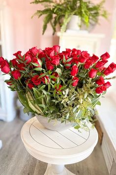 a white pedestal holding a bouquet of red roses on top of a wooden table next to a potted plant