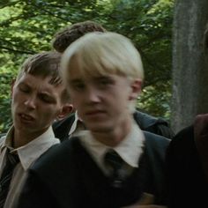 three young men standing next to each other in front of trees and bushes, one with blonde hair