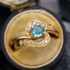 two gold rings with an aqua and white diamond center surrounded by small diamonds in a velvet ring box