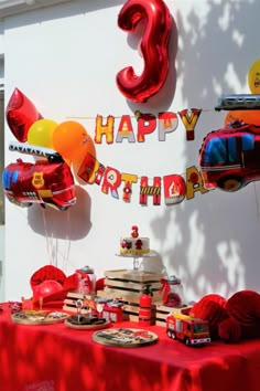 a red table topped with lots of cake and balloons next to a white wall that says 3 happy birthday