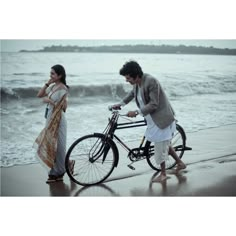 a man and woman standing next to a bike on the beach