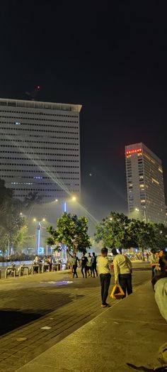 people are walking on the sidewalk in front of some tall buildings at night with bright lights