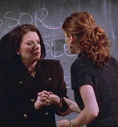 two women standing in front of a chalkboard with writing on it and one holding her hands together