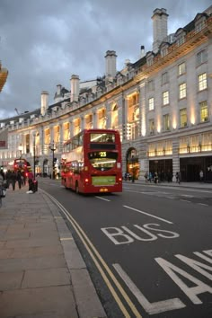 a red double decker bus driving down a street next to tall buildings with windows on each side