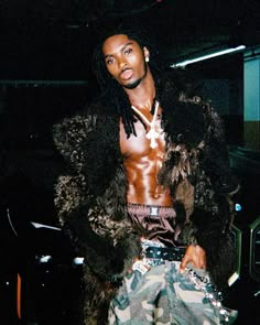 a man with dreadlocks standing in front of a car wearing camo pants