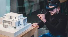 a man sitting at a table with a model house on it's side and a blind folded over his face