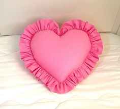 a pink heart shaped pillow sitting on top of a white bed with ruffled edges