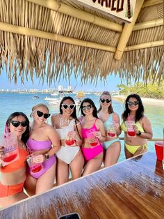 four women in bathing suits posing for a photo at the beach with drinks and straw umbrellas