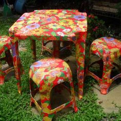 three colorful tables and stools sitting on the grass in front of some bushes with flowers painted on them