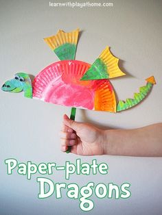 a hand holding a paper plate fish on top of a white wall next to a green plant