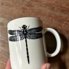 a hand holding a coffee mug with a dragonfly on it