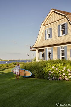 a man holding a surfboard in front of a house with flowers and bushes around it