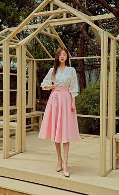 a woman standing in front of a wooden structure wearing a white blouse and pink skirt