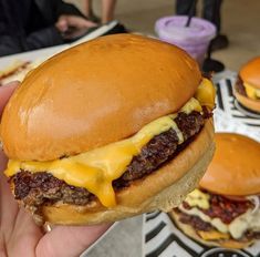 a hand holding a cheeseburger with other burgers in the background