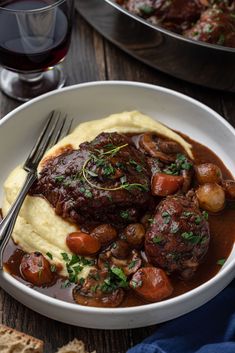 a white plate topped with meat and potatoes covered in gravy next to a glass of wine