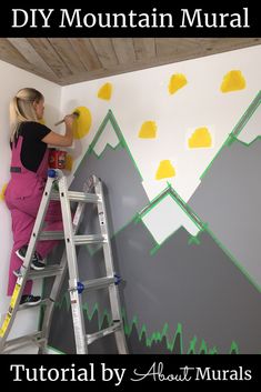 a woman is painting a mountain scene on the wall with yellow and green paintbrushes