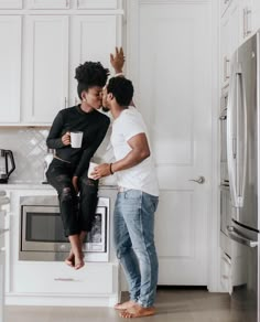 a man and woman are kissing in the kitchen next to an oven with white cabinets