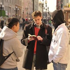 three men are standing on the street and one is looking at his phone while another man in a hoodie stands next to him