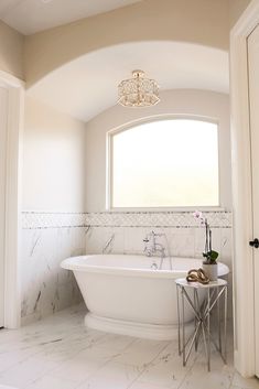 a white bath tub sitting under a window next to a table with a plant on it