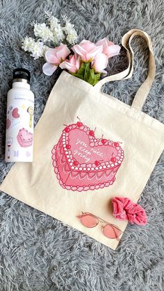 a tote bag with a pink heart on it next to sunglasses, flowers and a water bottle