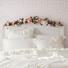 a white bed topped with lots of pillows next to a pink wall mounted flower crown