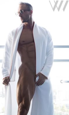 a man with no shirt standing in front of a window wearing glasses and a robe