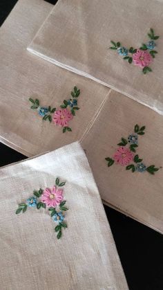 four embroidered napkins with pink and blue flowers on white linen, sitting on a black surface