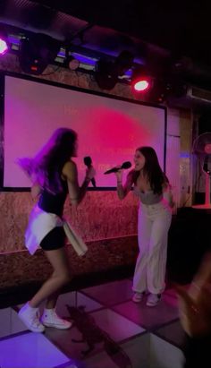 two women singing into microphones in front of a large screen with pink lights on it