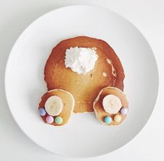 a white plate topped with pancakes covered in icing and candies next to a slice of banana