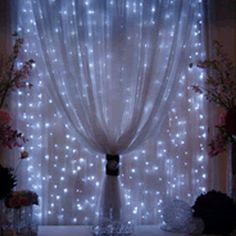 the curtains are covered with white lights in front of flowers and vases on display