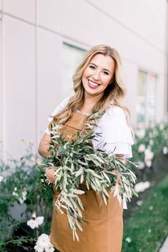 a woman in an apron is holding some plants and smiling at the camera while standing outside