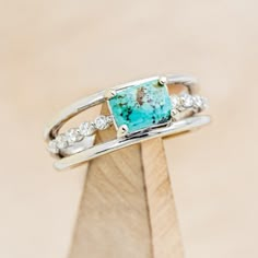 a ring with a turquoise stone and white diamonds on top of a wooden stand in front of a beige background