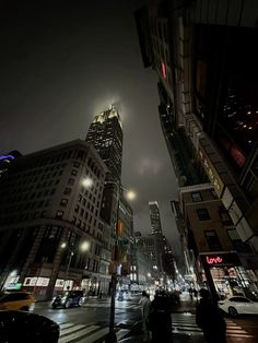 people are walking down the street at night in new york city, with skyscrapers lit up behind them