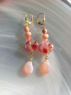 Earrings with handcrafted oval glass flower beads in a golden flower dish, hanging pink glass drops, Cherry Quartz, Pink Opal and Aventurine beautifully embellish these earrings. Very beautiful oval beads measuring 19mm wide, 12mm high and 6mm thick. They are handmade by an artist from Canada in New Brunswick. They are embellished with 6 mm Cherry Quartz, 8 mm x 5 mm Peruvian Pink Opal rounds and 5 mm Aventurines. Gold plated shell and lever back ear hooks. These earrings measure 8cm or 2 and a quarter inches from top to bottom. Each of these loops weighs 6 g. Handmade Pink Flower Earrings With Round Beads, Pink Handmade Flower Drop Earrings, Handmade Pink Oval Beads Jewelry, Pink Dangling Bead Flower Earrings, Pink Czech Glass Dangle Earrings, Golden Flower, Ear Hook, Pink Opal, Pink Glass