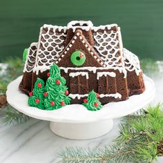 a gingerbread house decorated with icing on a white cake platter surrounded by evergreens