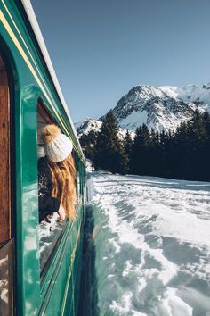 a woman looking out the window of a train on snow covered ground with mountains in the background