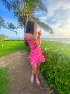 a woman in a pink dress drinking from a glass next to the ocean and palm trees