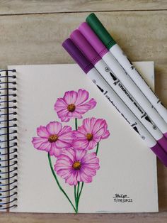 three markers are sitting on top of a notebook with some flowers painted on it and four pens in front of them