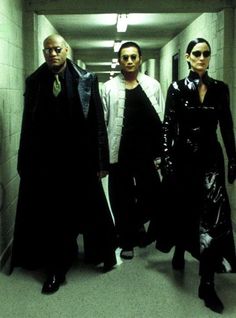 three people walking down a long hallway in black clothes and leather jackets, all looking at the camera