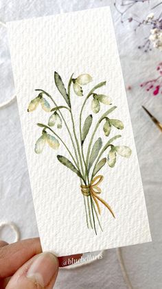 a hand holding up a card with watercolor flowers on it and thread in the background