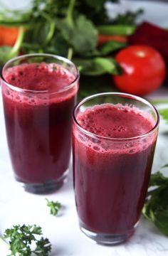 two glasses filled with red liquid sitting on top of a white table next to vegetables