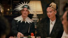 two people sitting at a table with plates and glasses in front of them, one wearing a headdress