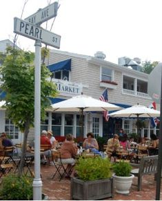 people are sitting at tables outside in front of a restaurant with umbrellas and potted plants