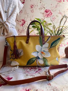 This Magnolia Buddy Bag is a luxurious accessory designed to elevate your daily look. Crafted from a mustard yellow fabric adorned with beautiful large magnolias, this bag is sure to add an artistic, elegant touch to any outfit. The included tassel adds a stylish, sophisticated detail. This chic sack will be your new BFF! Get a grip on its leather handles, and you can easily adjust the strap to your comfy fit. It boasts a water-repellent taffeta lining, a nifty zippered pocket, and even a split Knitting Needle Storage, Boho Chic Bags, Handcrafted Handbags, Local Yarn Shop, Yarn Storage, Project Bags, Bag Obsession, Yarn Bag, Boho Bags