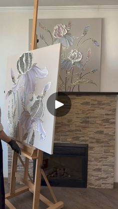 a woman standing in front of an easel painting flowers on a white canvas with a fireplace behind her