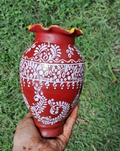 a hand holding a red vase with white designs on it in front of green grass