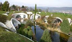 an unusual house on top of a hill next to a pond and forest area with people sitting in it