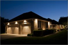 two garage doors are lit up at night in front of a house with bushes and shrubs